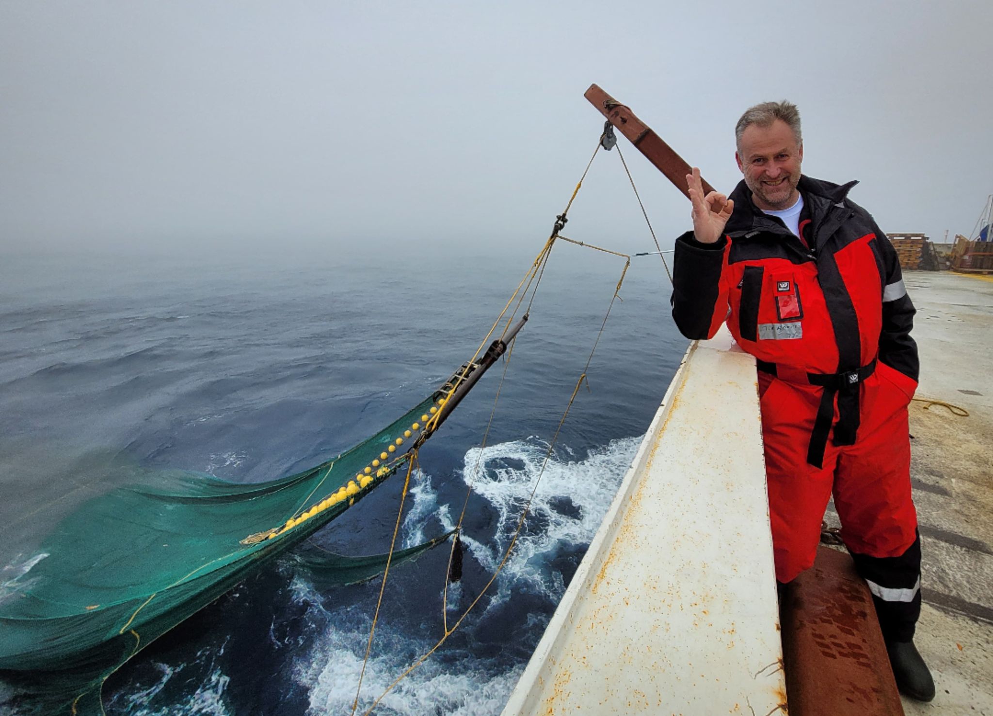 mann på skip med tommel opp, trål hengende ut i havet bak