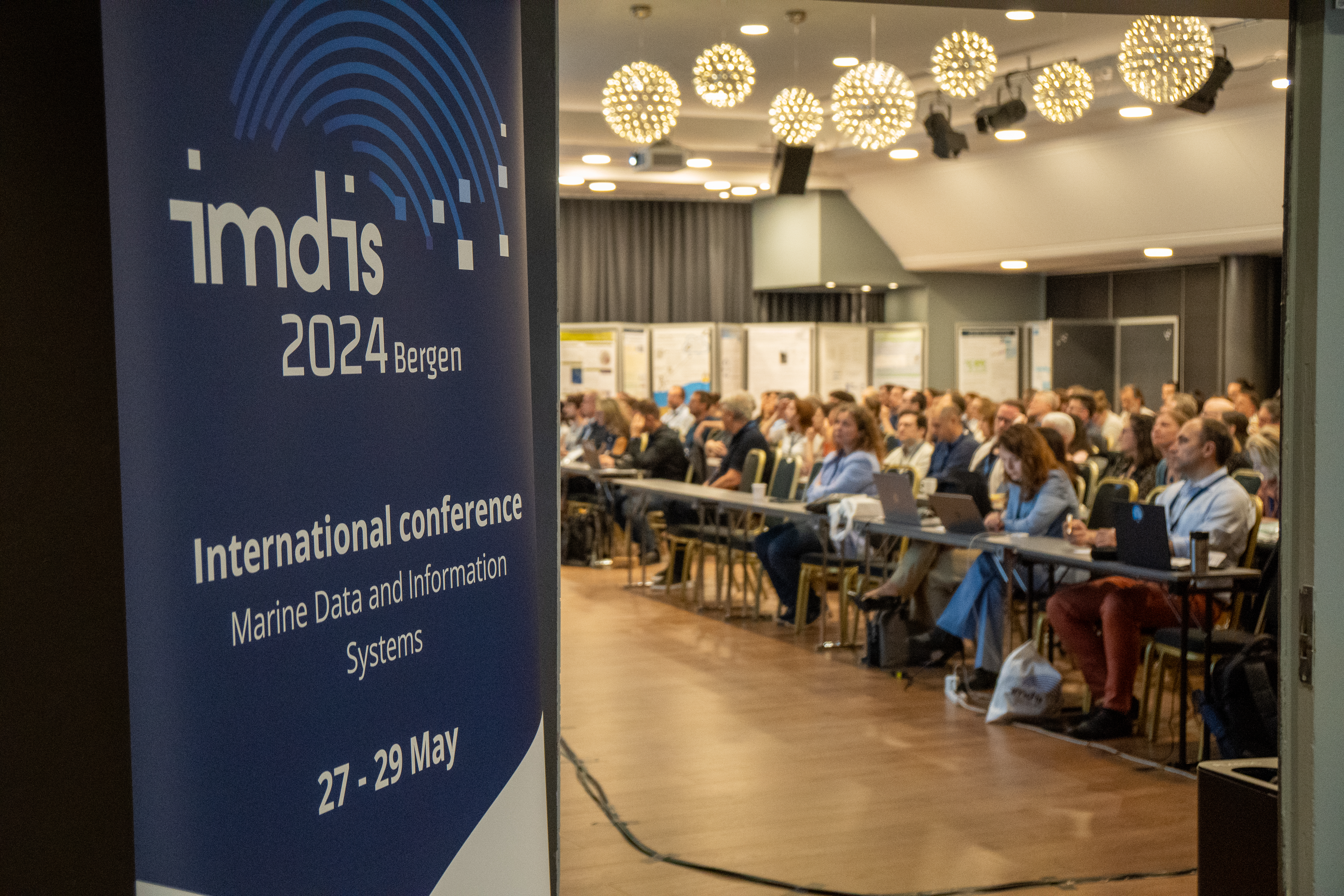 På en plakat til venstre står IMDIS. Til høyre gjennom en døråpning, sitter mange folk og lytter til et foredrag