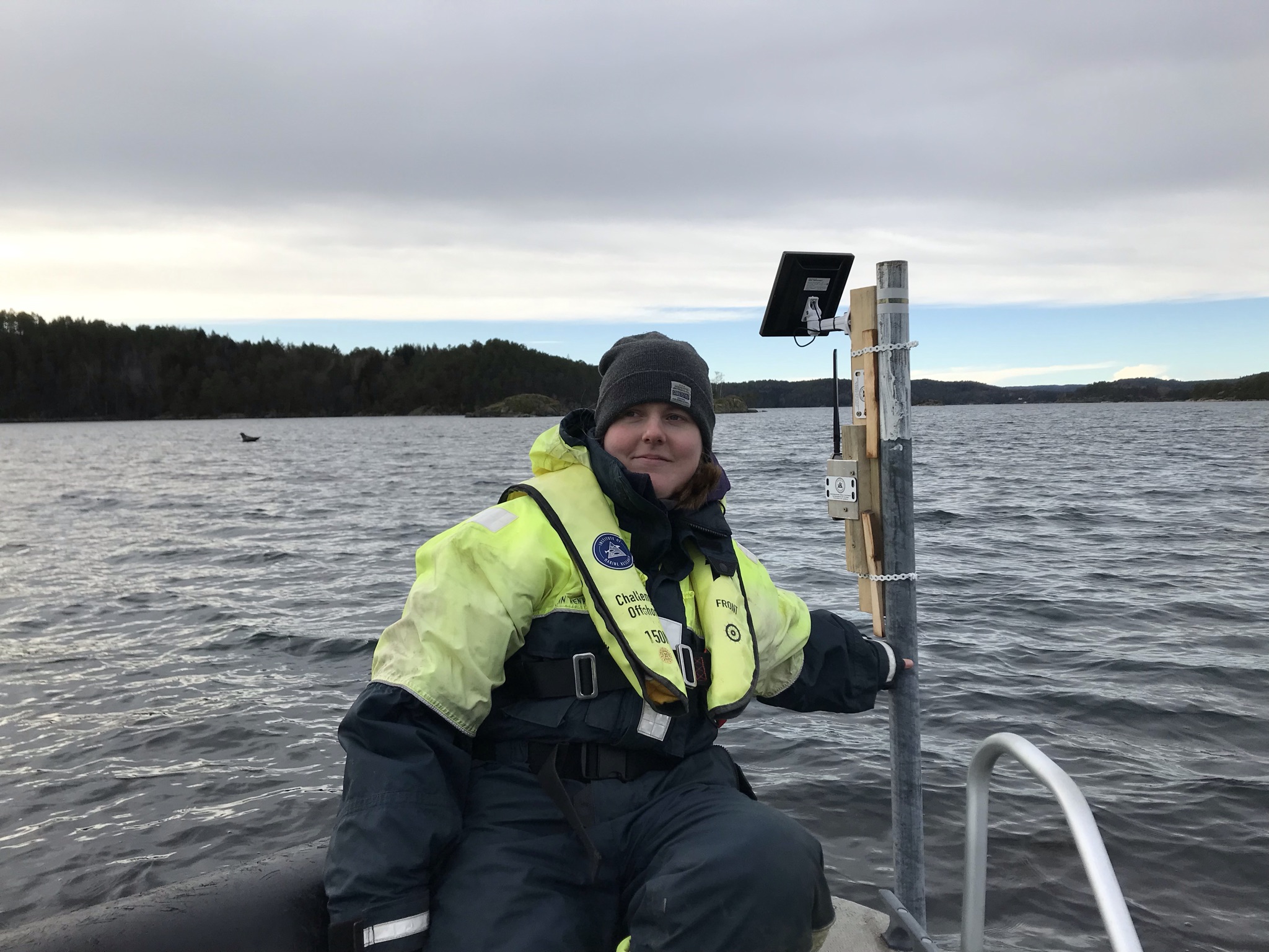 En kvinne sitter i en båt på havet, bak henne står en stolpe. På toppen av stolpen henger et viltkamera