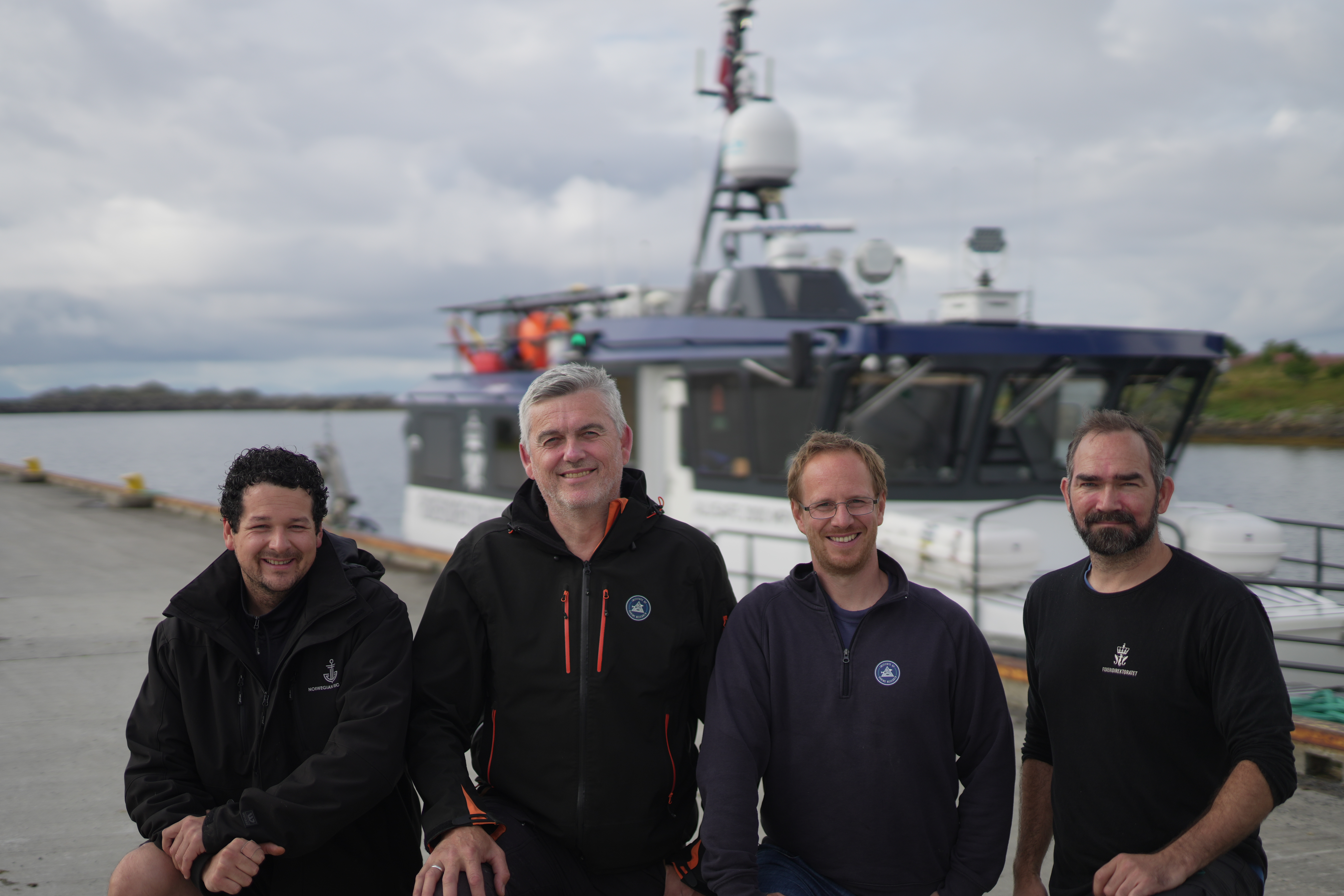fire menn smiler til kamera, stående på en kai, med en båt i bakgrunnen