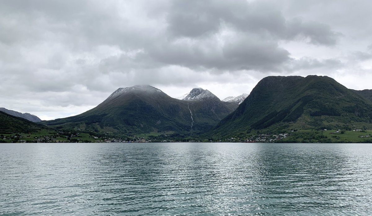 
Grønt vann i fjorden i bakgrunnen, fjell med bebyggelse i en vik i bakgrunnen.