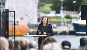 Kvinne på podium med mennesker i forgrunnen og bybilde med to båter og en eldre steinbygning (Håkonshallen) i bakgrunnen.