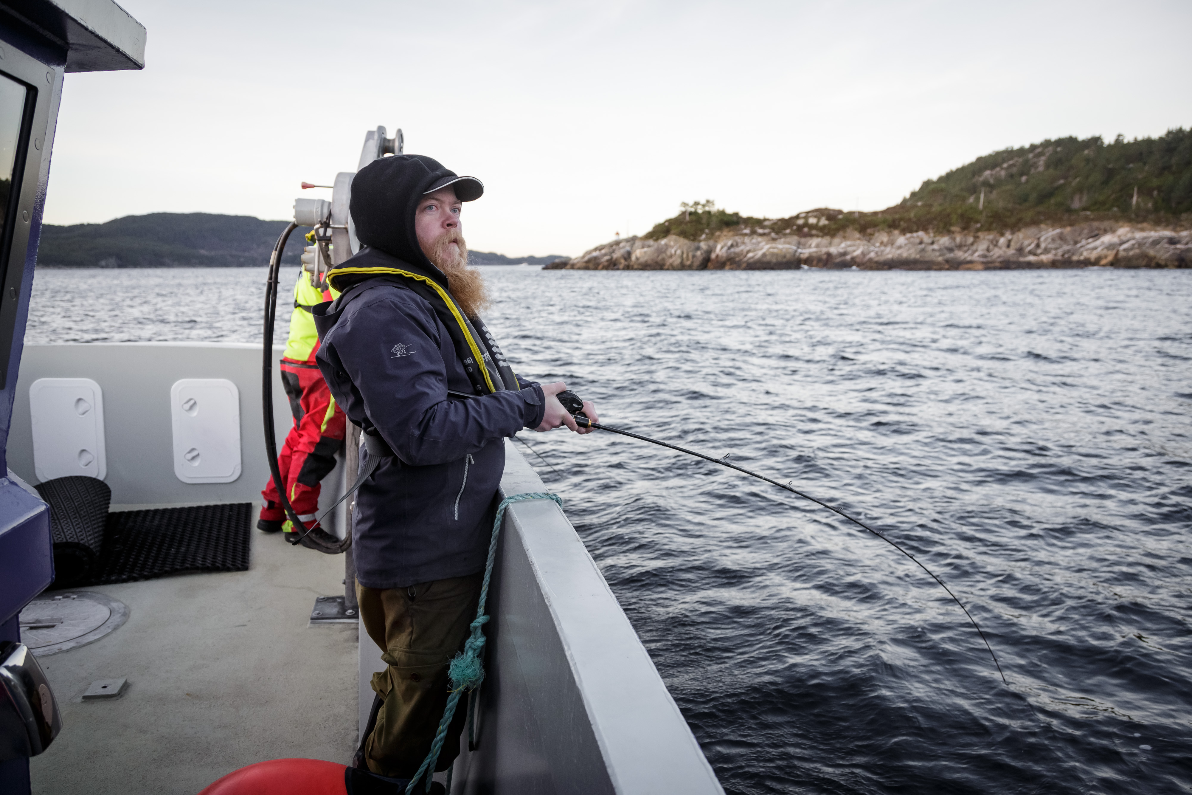 Mann med blå caps og langt skjegg står i båt med fiskestand over rekka og fisker.