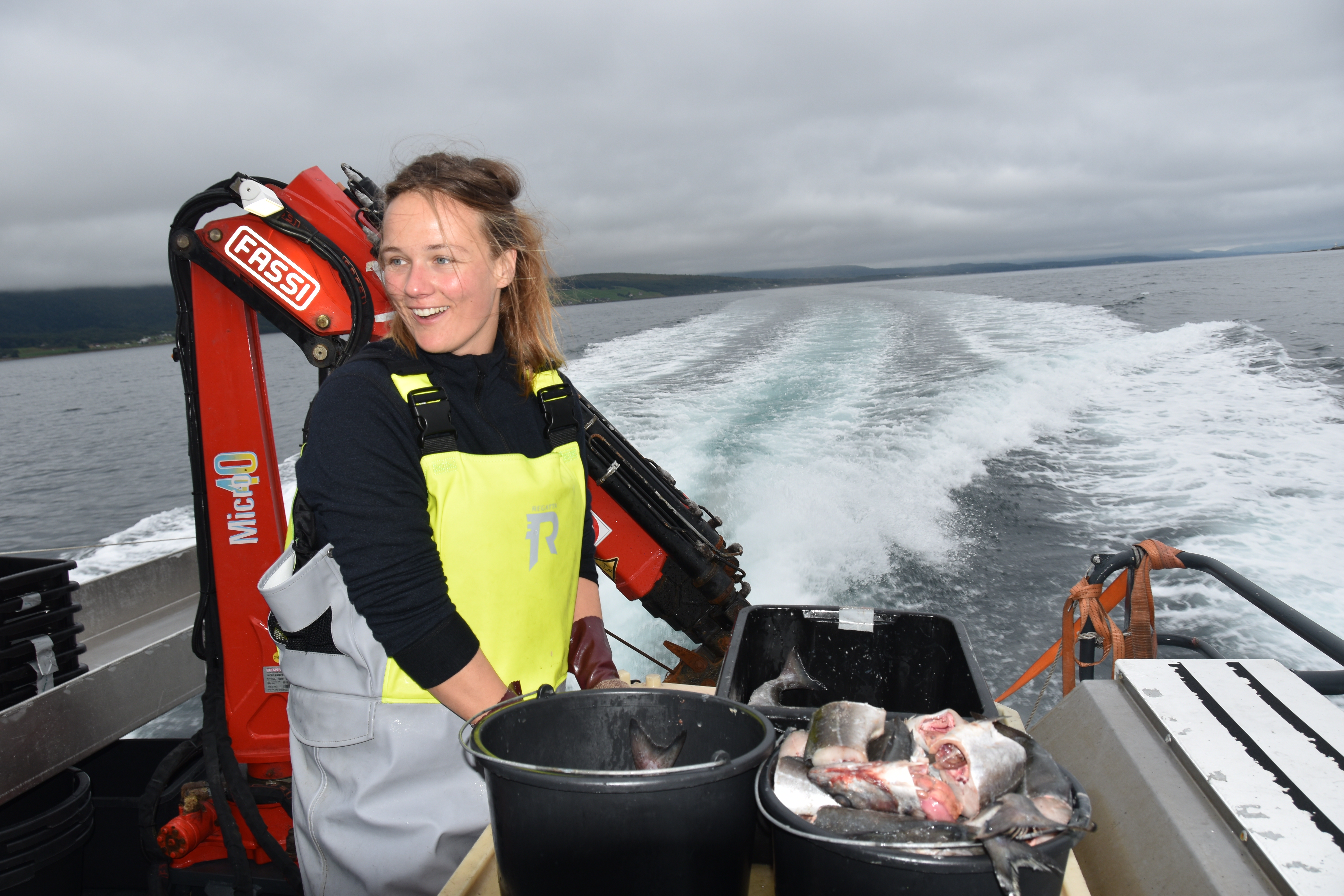 Stipendiat Johanna gjør klart for å pakke agnposer. Foran henne står en bøtte med oppkuttet fisk. Hun smiler, og man kan se kjølvannet etter båten i bakgrunnen. 
