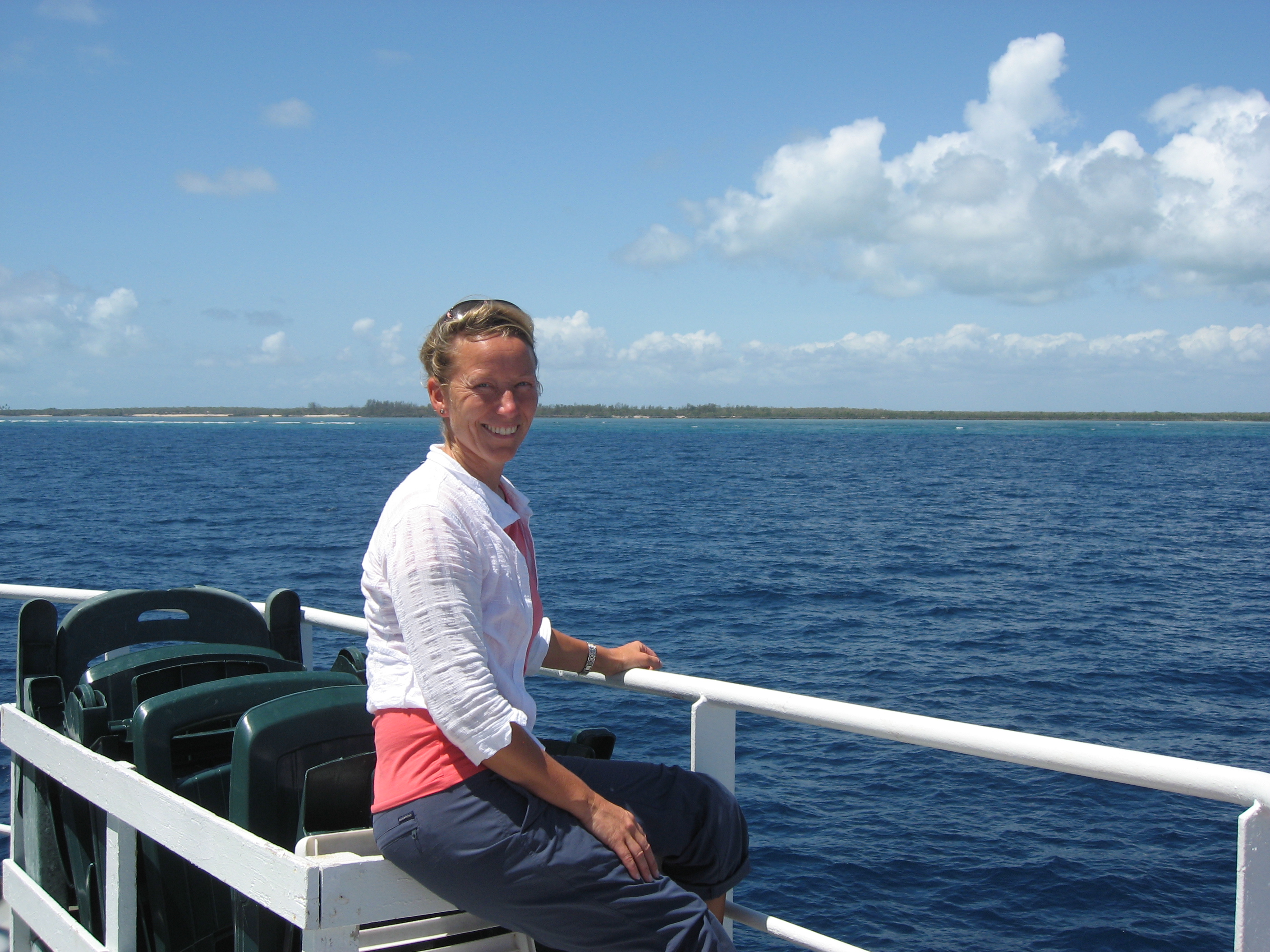 Kvinne på dekk på et skig med rolig sjø og flattliggende land og strand i bakgrunnen
