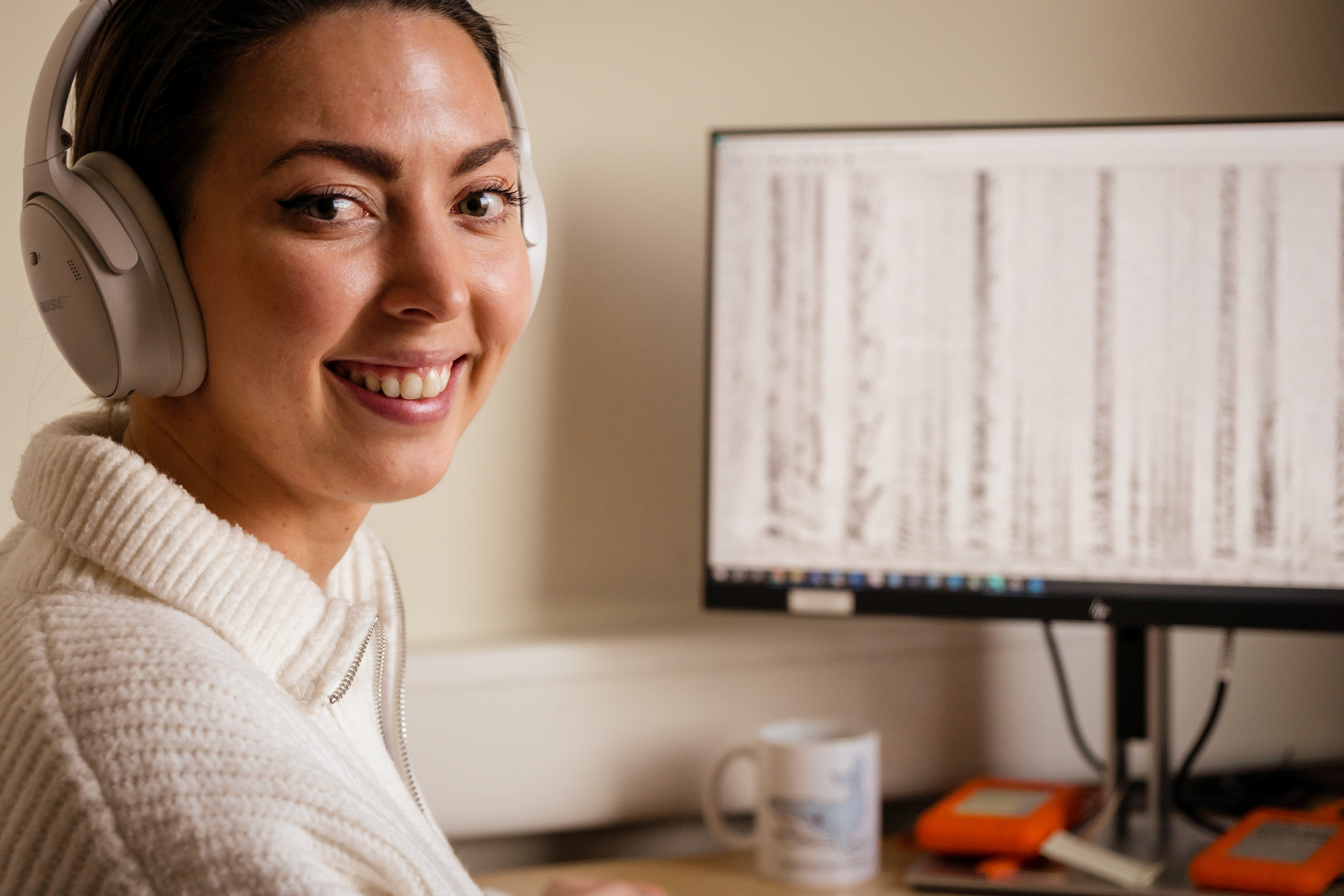 Smilende ung kvinne med øretelefoner på hodet ser inn i kamera. Bak henne er en PC. 