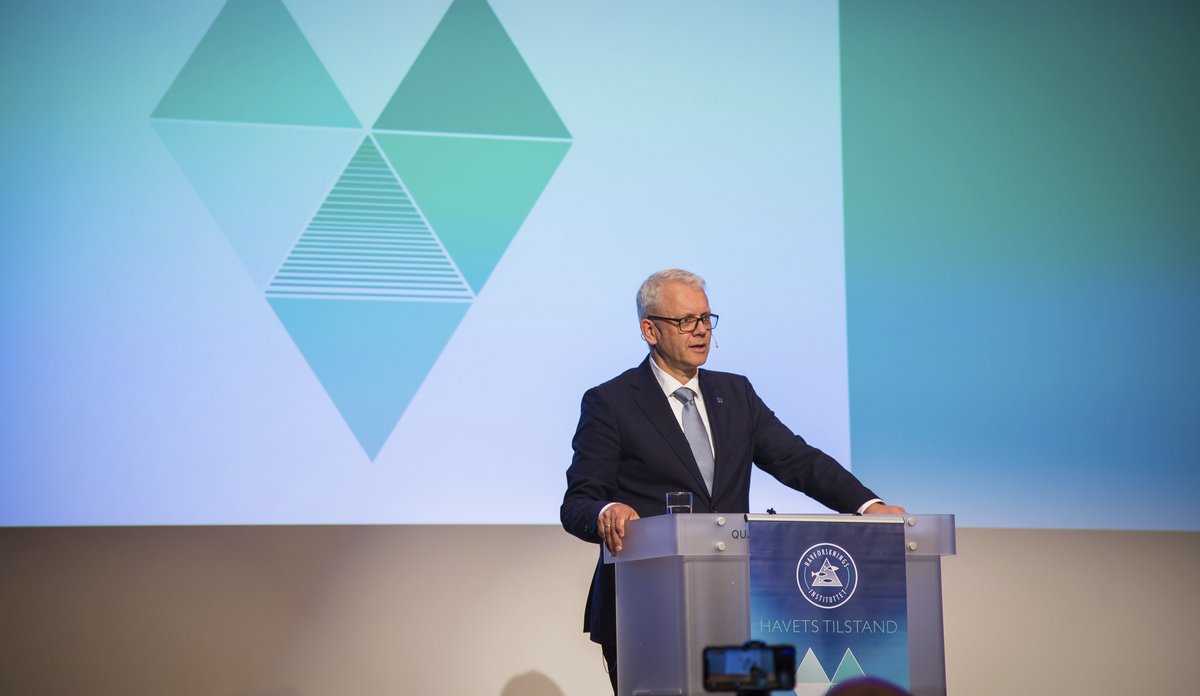 
mann står bak talerstol på lyssatt scene med hjerteformet logo på skjerm bak seg 