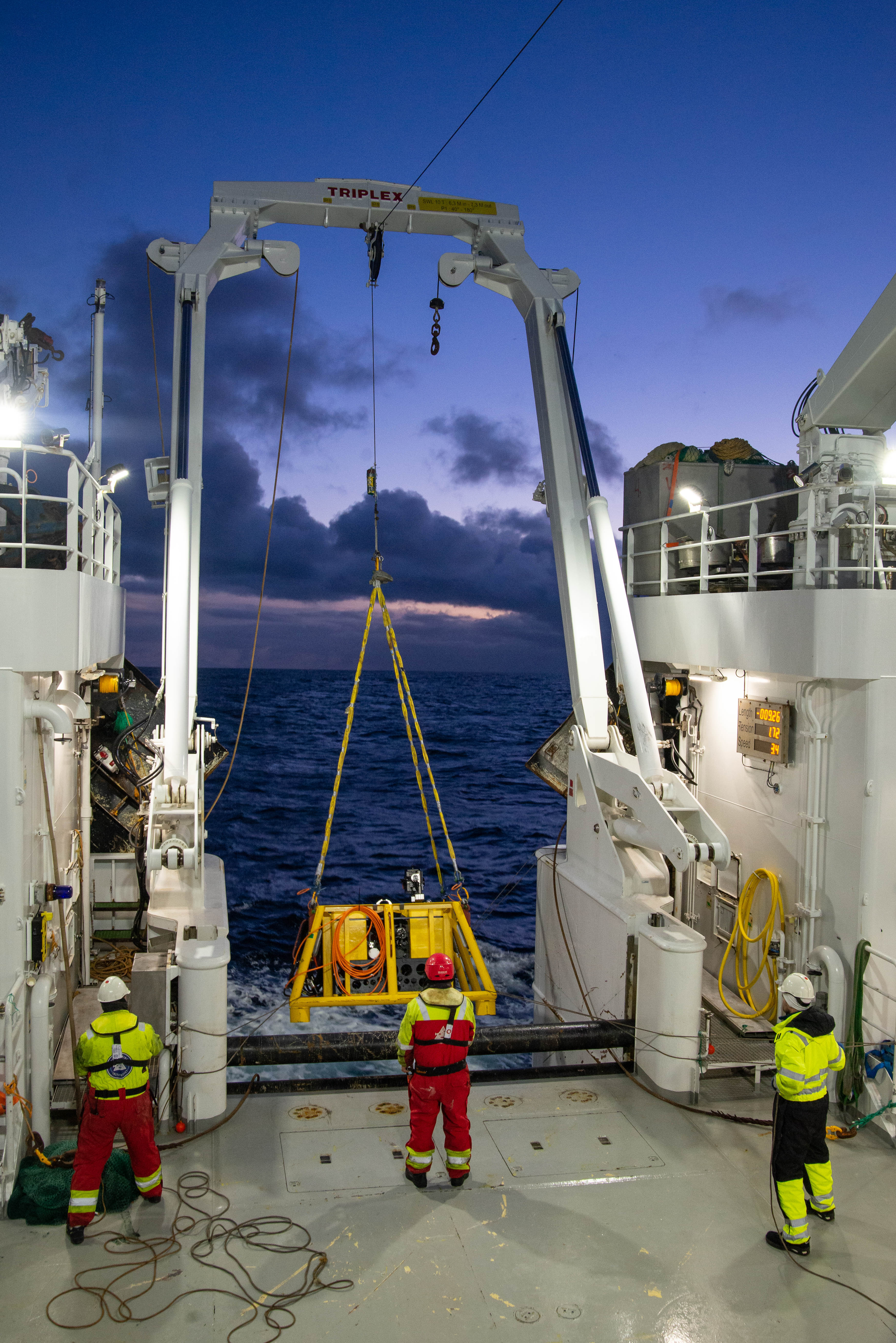 Tre mannskap, sett bakfra, står på dekk og gjør klart for å senke utstyr ned i sjøen.