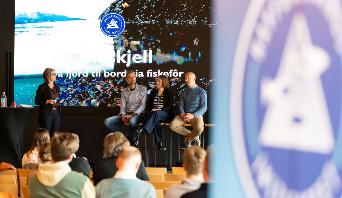 
Bilde fra et arragement. På scenen sitter tre paneldeltagere på hver sin stol. Ordstyrer står