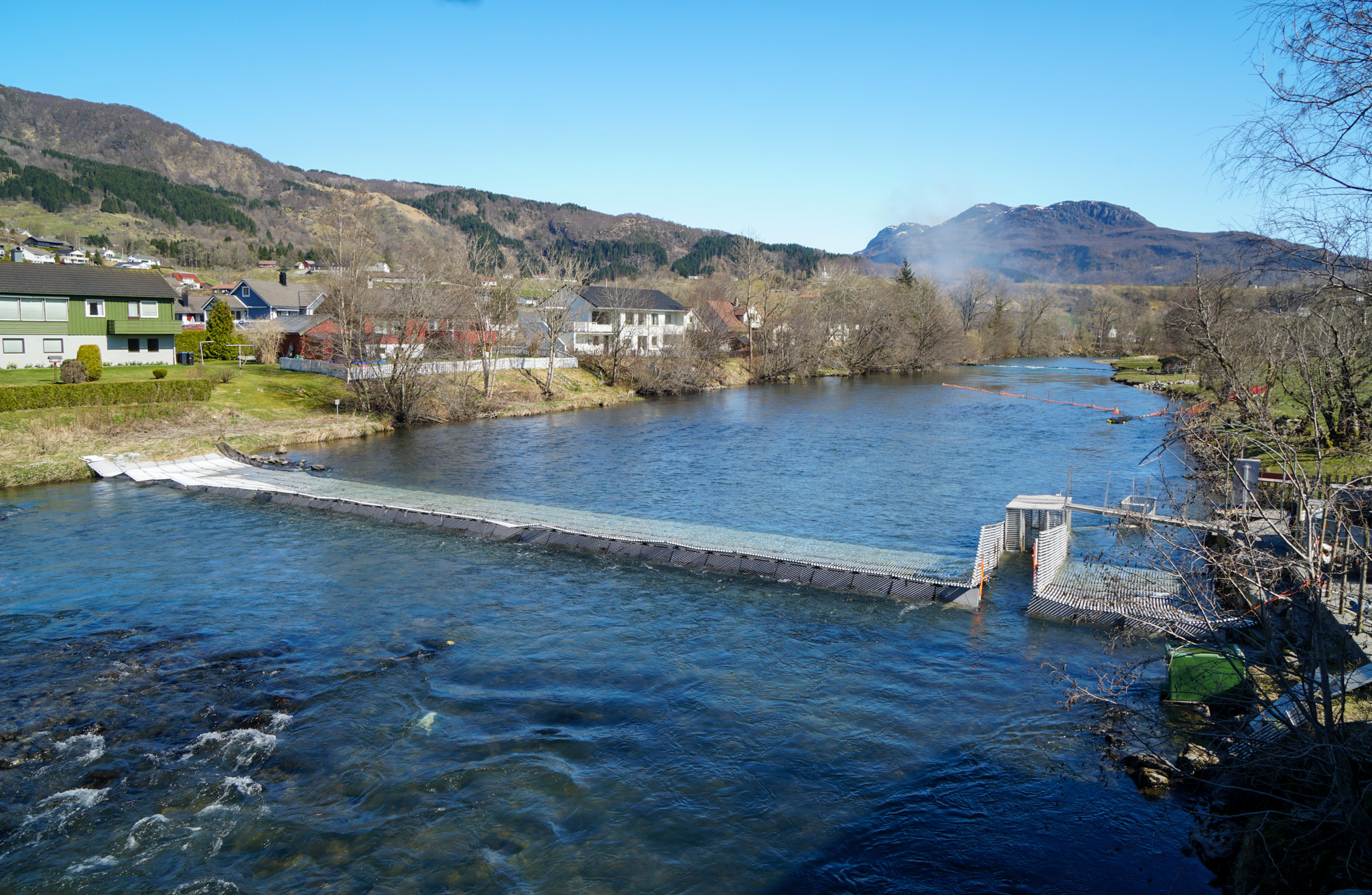 Elv med ein flyteristfelle på tvers. Bakkar og hus i bakgrunnen.