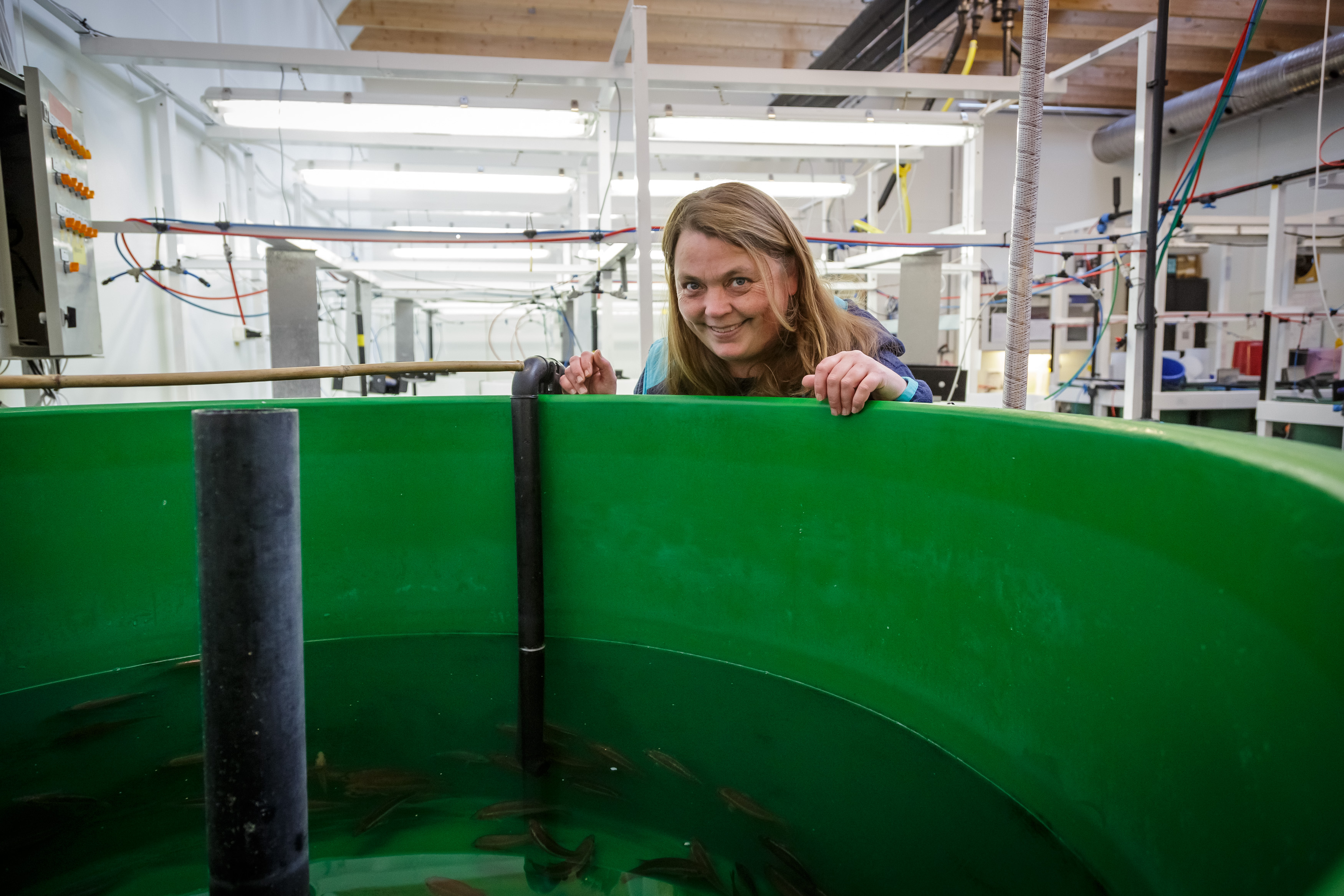 Smilende kvinne står bøyd over et stort, grønt kar inne i en hall med mange rør og ledninger i bakgrunnen.