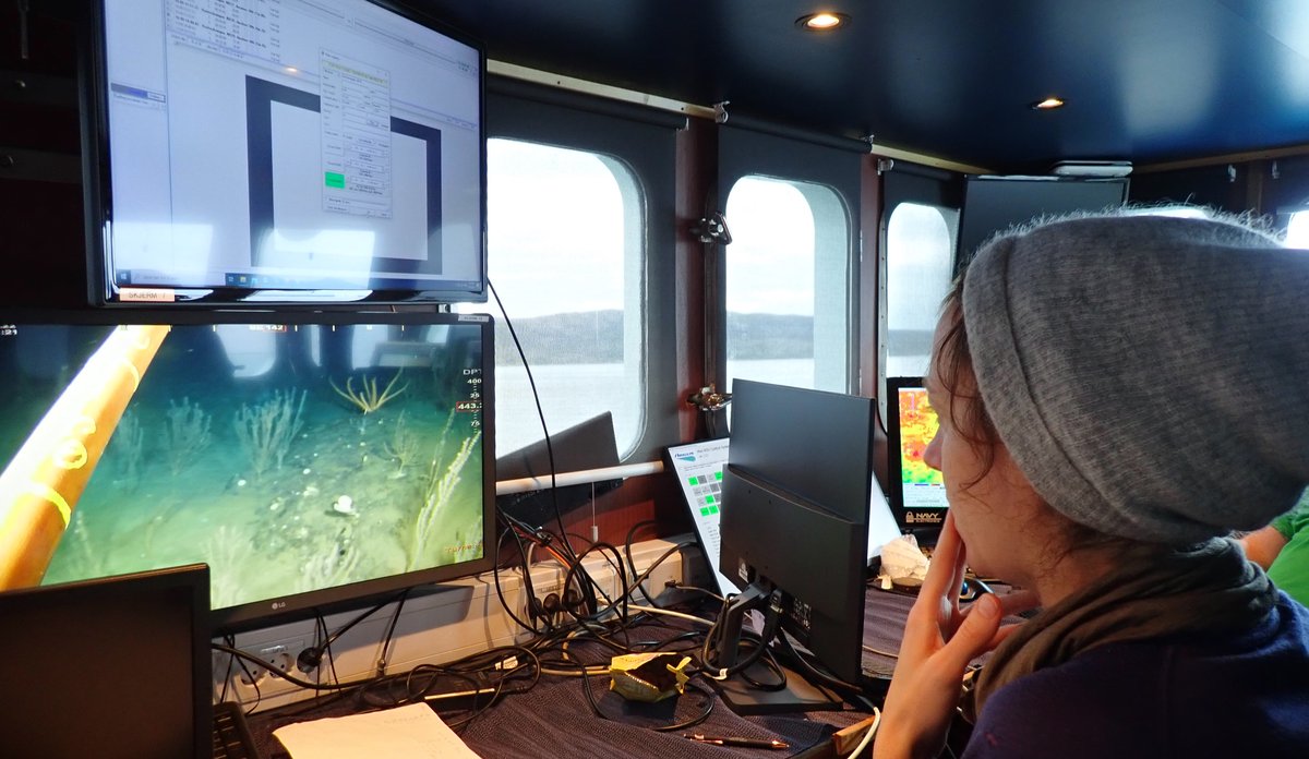 
Ei kvinne med grå strikkahue sit i eit kontrollrom på eit fartøy. Ho ser på to dataskjermar framfor seg, som viser kva som er på havbotnen