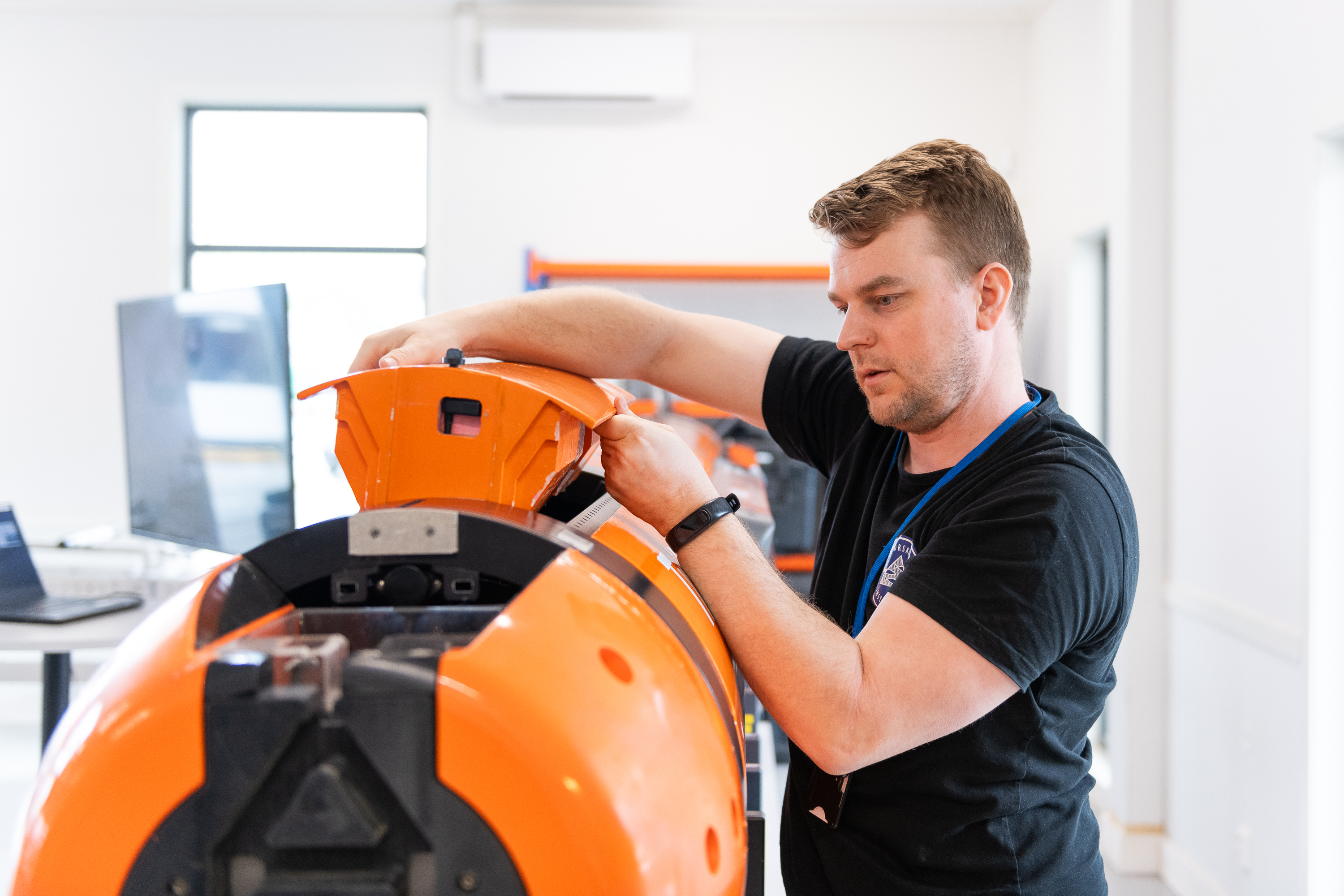 Mann i svart t-skjorte løfter et oransje deksel fra en stor, liggende sylinder som er svart og oransje. 