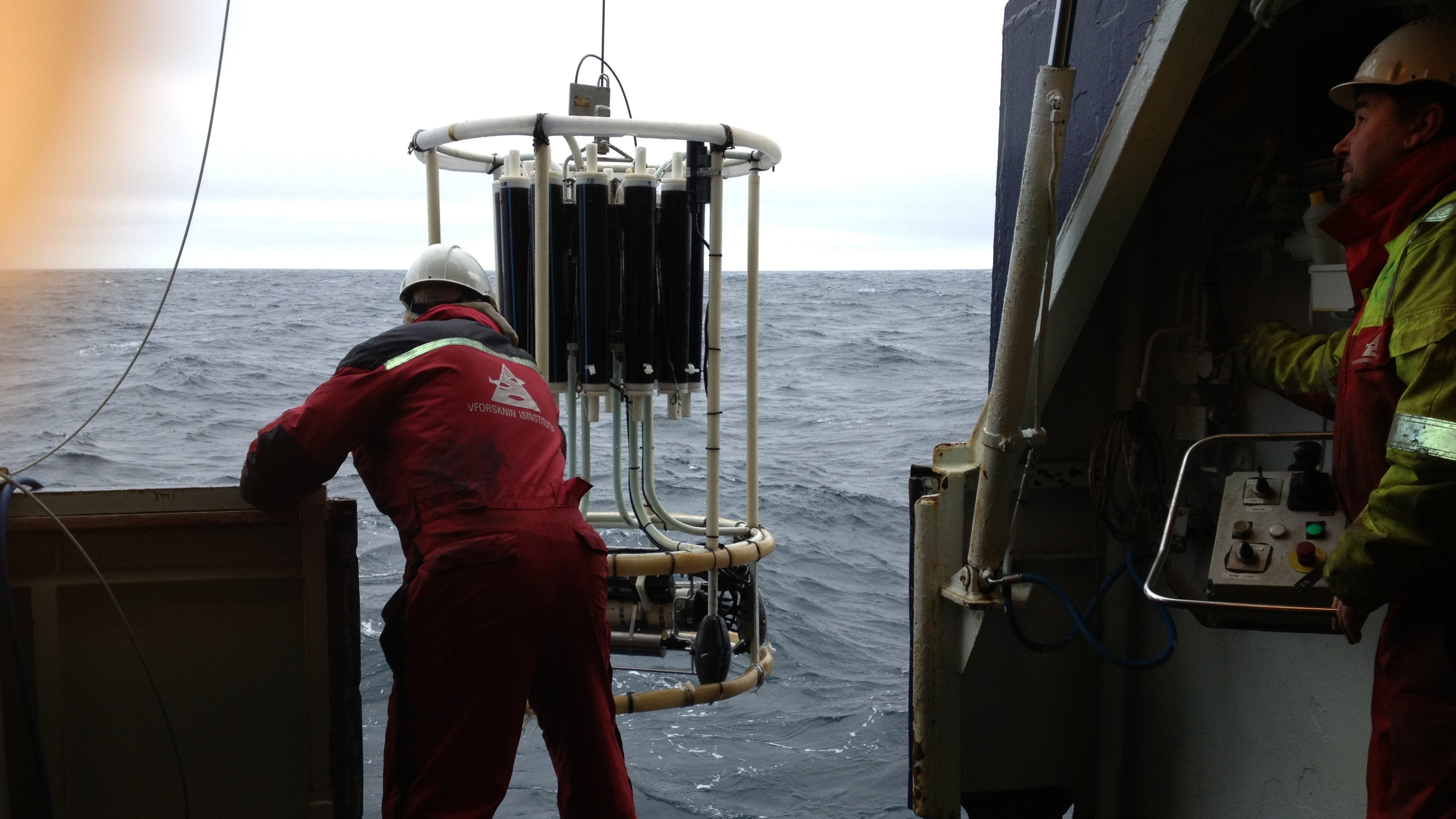 Vi ser ryggen på en havforsker som dytter en vannhenterkrans ut i havet, og fronten på en som styrer en vinsj.