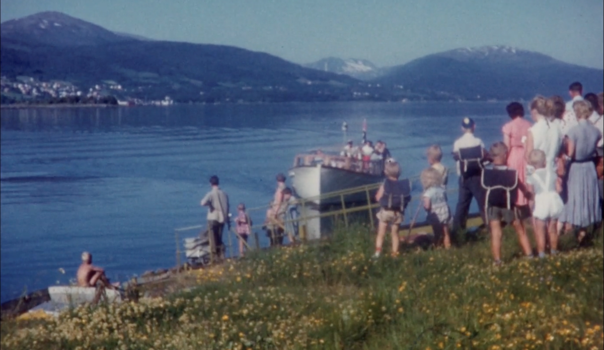 
en sommerkledd folkemengde står på land og venter på en båt 