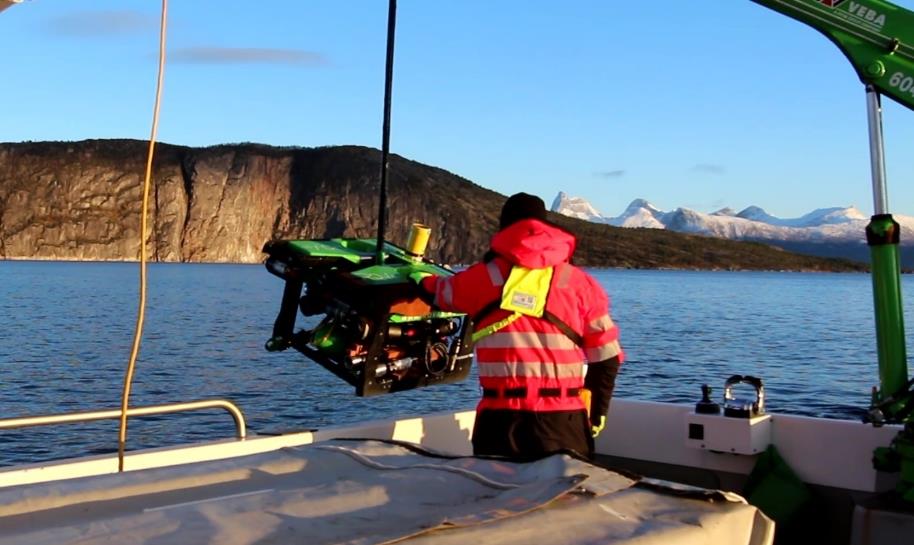  ROV henger fra kran og er på vei ned over ripa, toktpersonell står på dekk og holder på ROV. I bakgrunnen sees sjøen, og fjell som bader i vintersol.