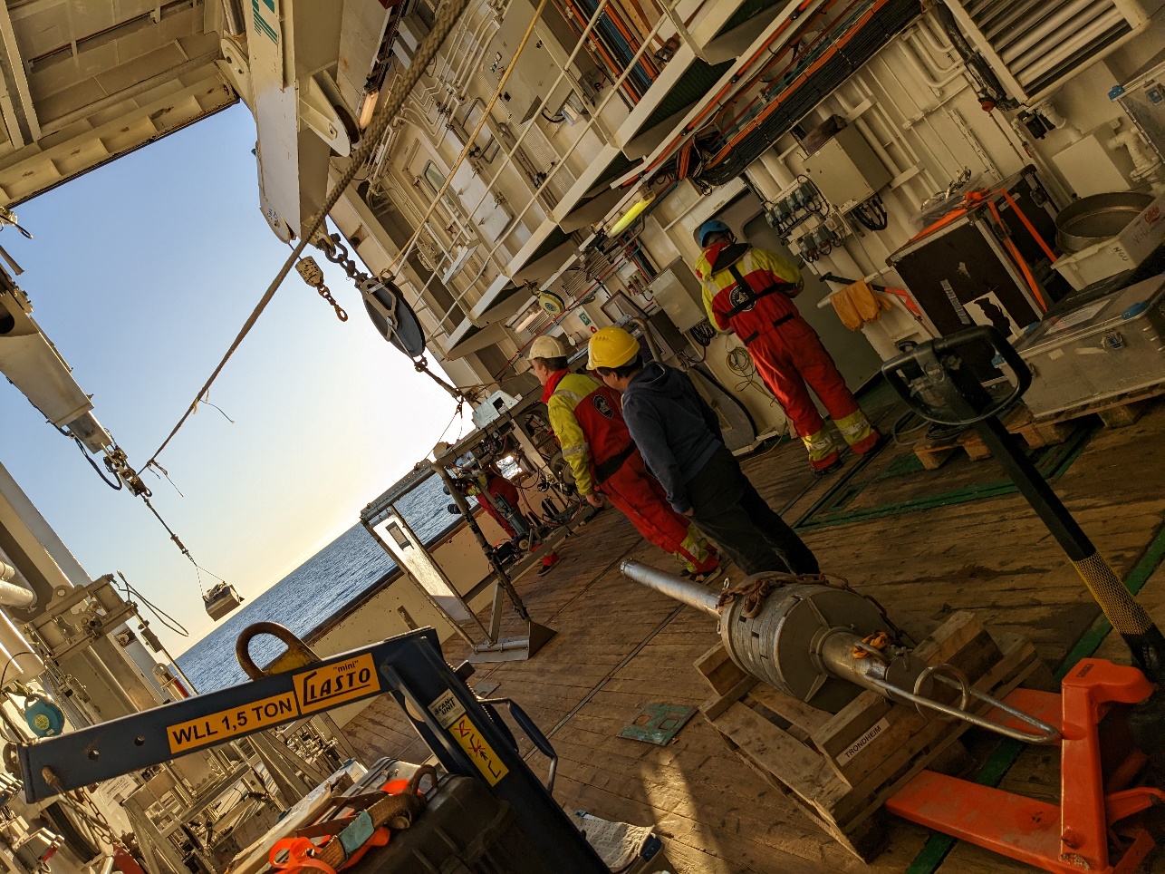 A photo with an oblique view through the hanger as 3 people look on with helmets on, the grab hangs from the winch wire while blue skies and calm seas can be seen outside.