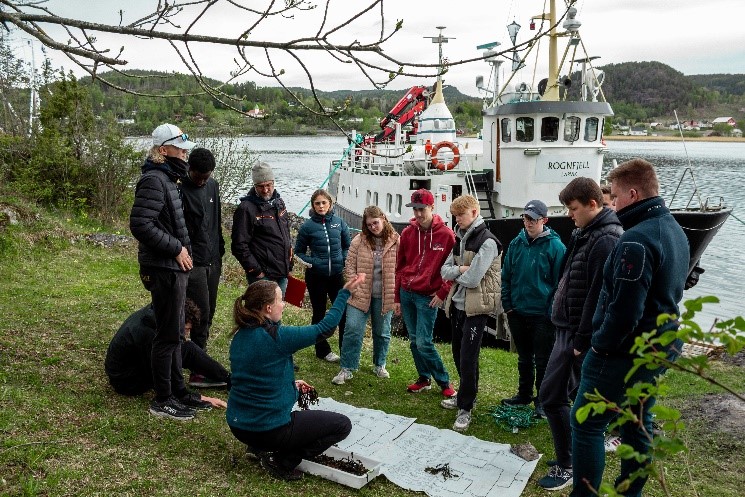 Bilde av elever som står på land med lærer i forgrunnen som viser fram en tang, og med skolebåten Rognfjell i bakgrunnen.