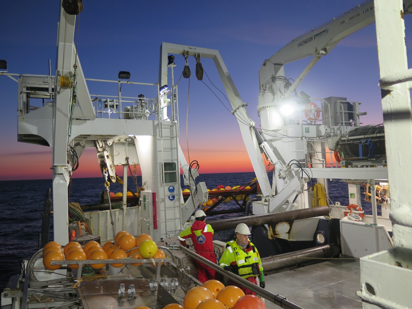 BIldet viser to medlemmer av mannskapet på dekk, deler av video-sleden og deler av trålen. Bildet er tatt ved solnedgang, og vi ser en oransje strime i horisonten. 