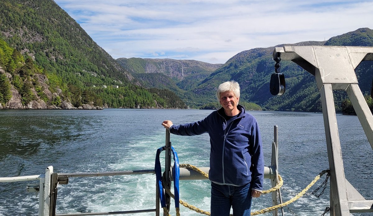 
Foto av en mann som står helt bak på en båt. Sjø og fjell i bakgrunnen.
