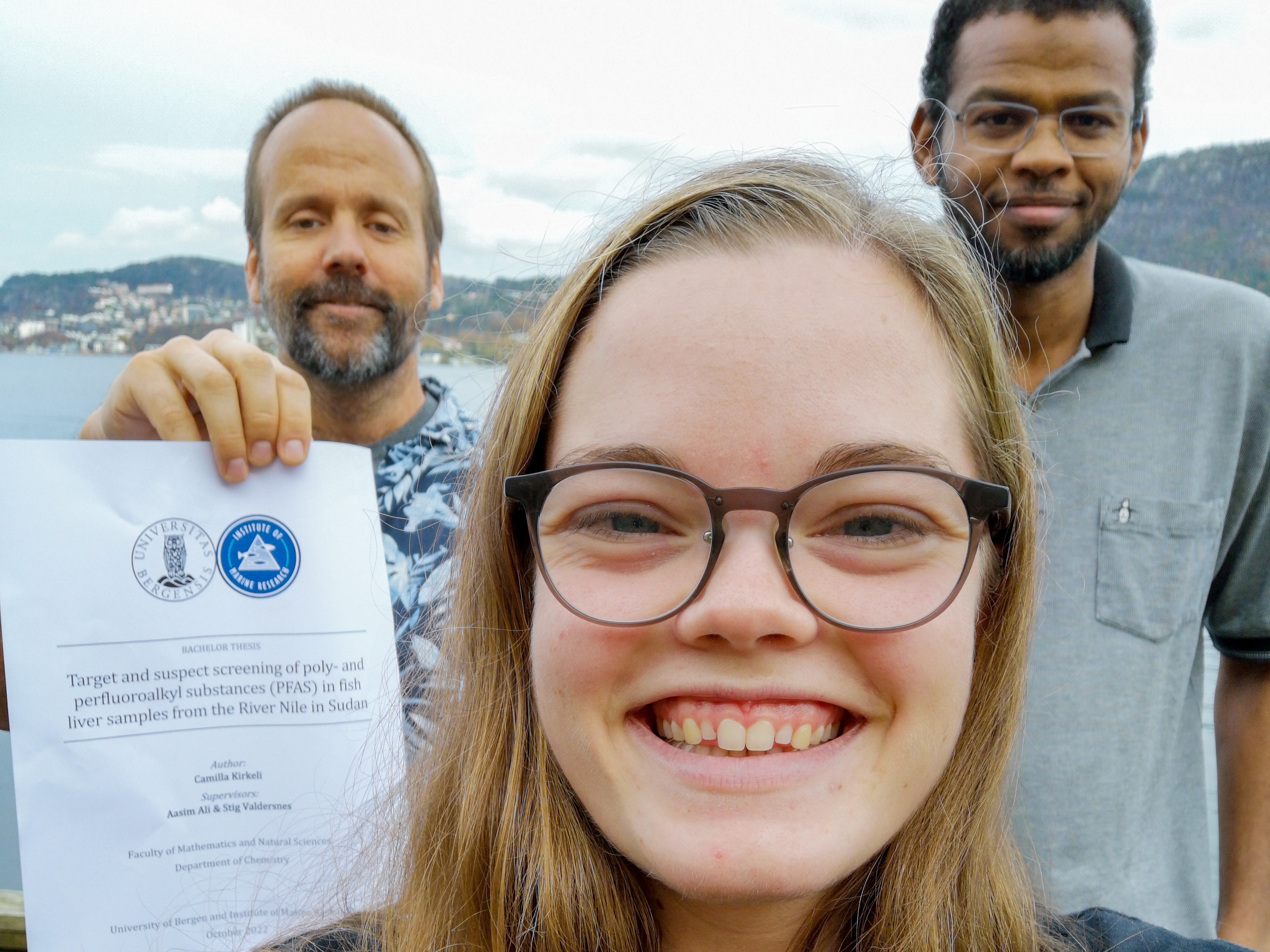 kvinne smiler i kamera med to menn bak, den ene holder opp en rapport 