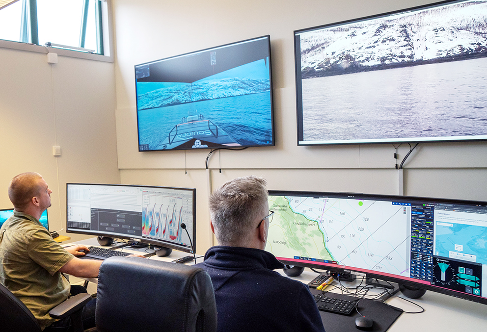 To menn sitter i operasjonssentralen Bergen og ser utsikten fra USVen i nord. De sitter på kontorstoler foran en rekke skjermer som viser området. 