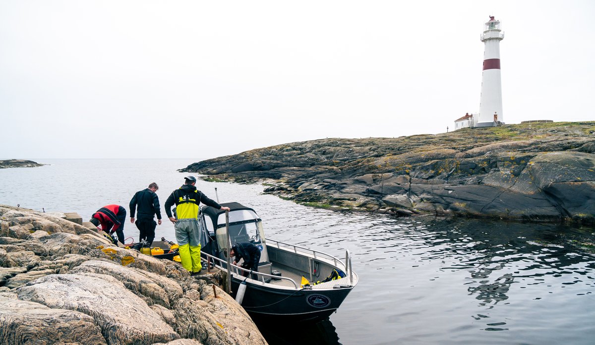 Mennesker i båt ved holme og fyrtårn.