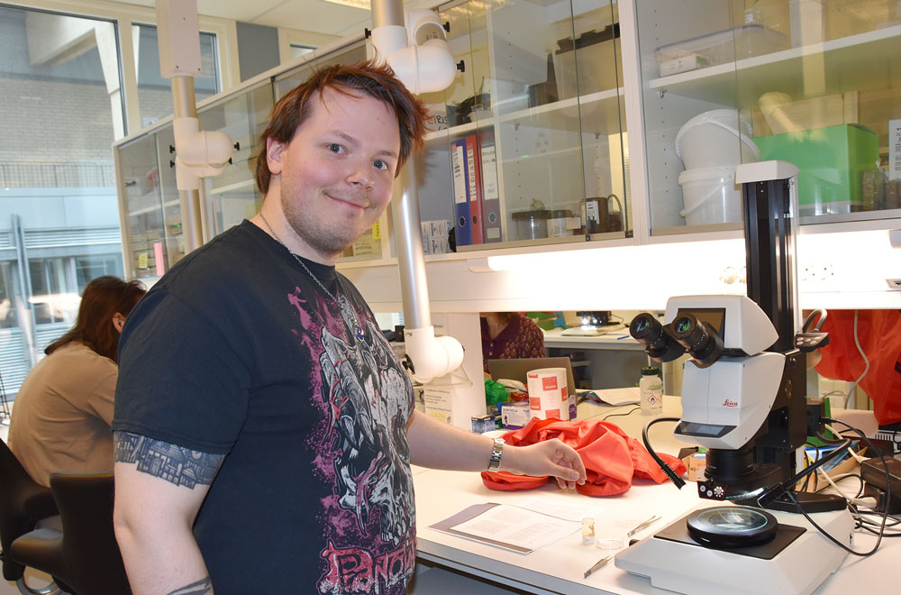 En mann står ved siden av et mikroskop. Han smiler og ser i kameraet.